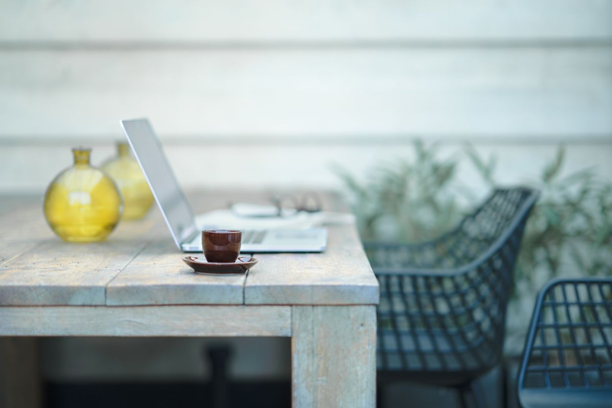 Laptop, teacup, and vases on a wooden table, with chairs and foliage in the background.