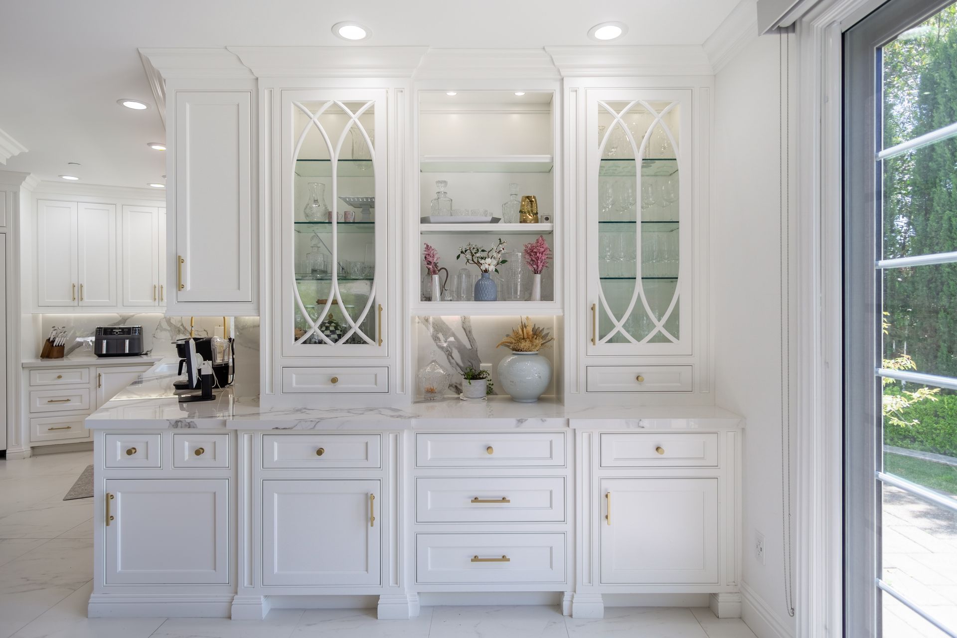 Bright white kitchen with glass-front cabinets, island, and large sliding doors opening to a green yard