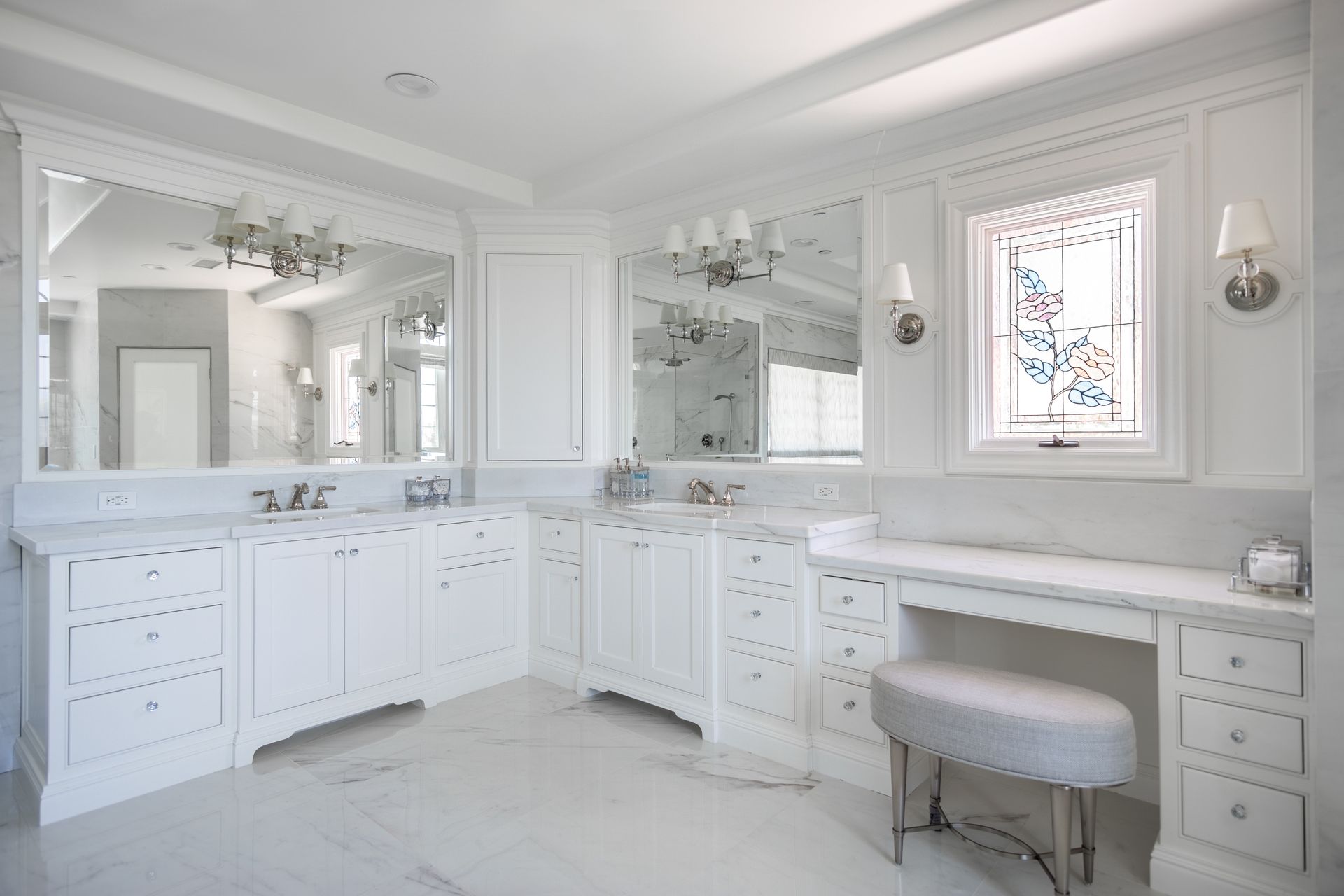 Bright white bathroom with double vanity, large mirrors, marble counters, and a cushioned stool