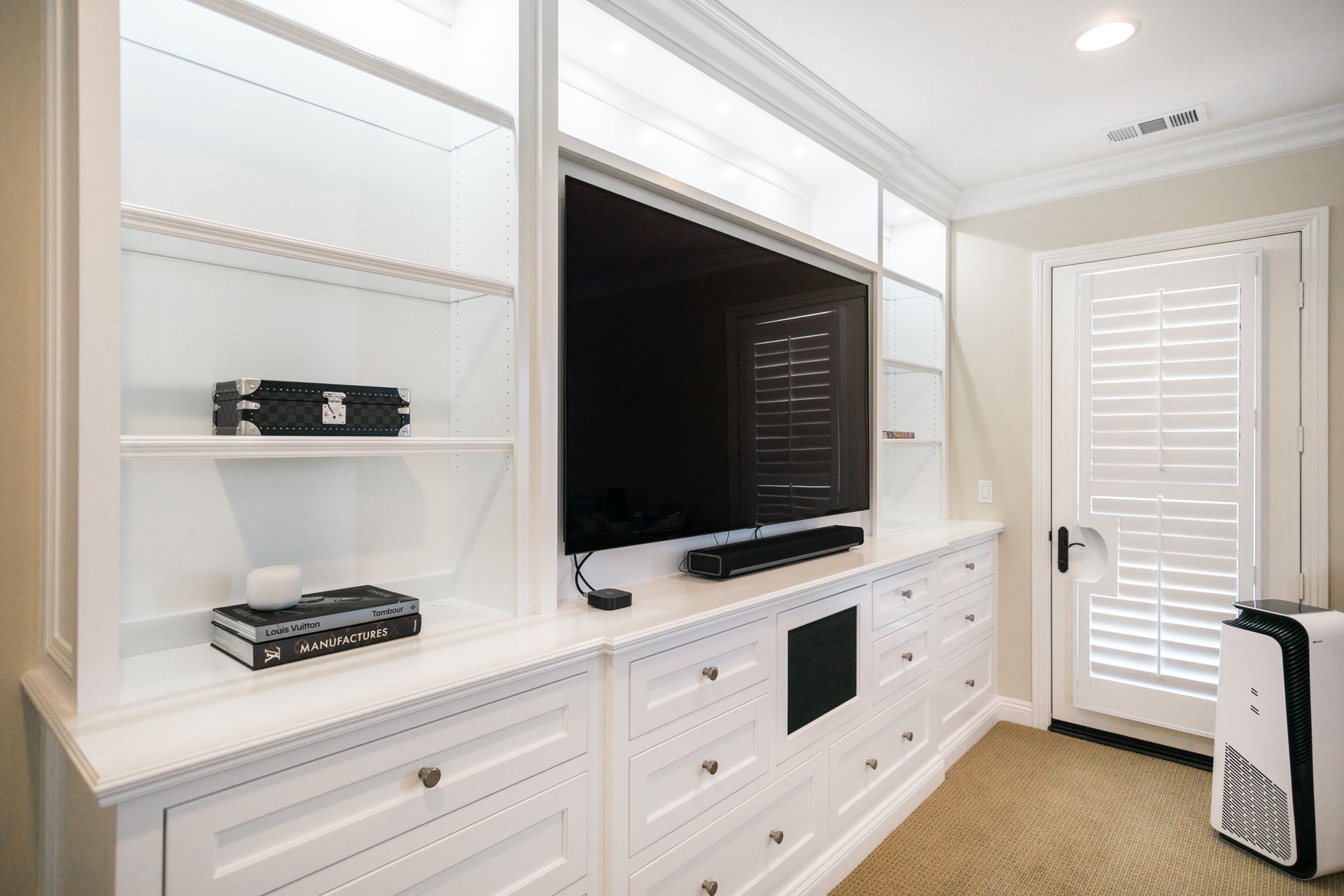 Built-in white media wall with large TV, cabinets, shelves, and a luggage bag in a bright room