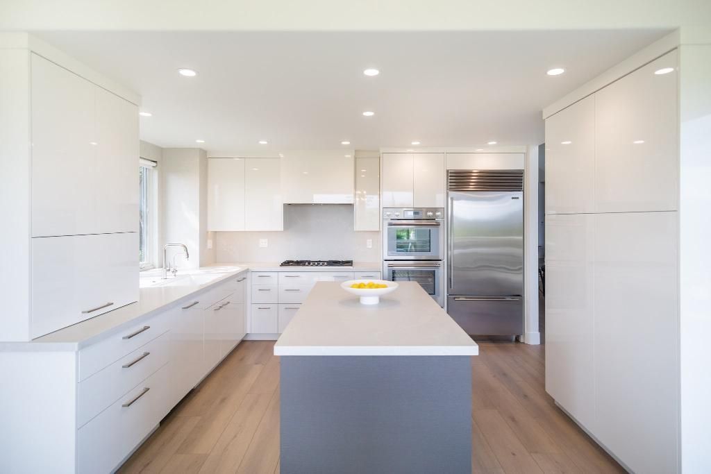 Bright modern white kitchen with a central island, wood floors, and stainless steel refrigerator.