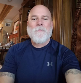 Smiling bald man with a white beard in a navy shirt, seated indoors with wood-paneled walls.