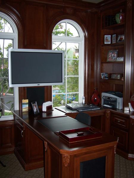 Wood-paneled home office with a large monitor desk, windows, shelves, and a printer nook