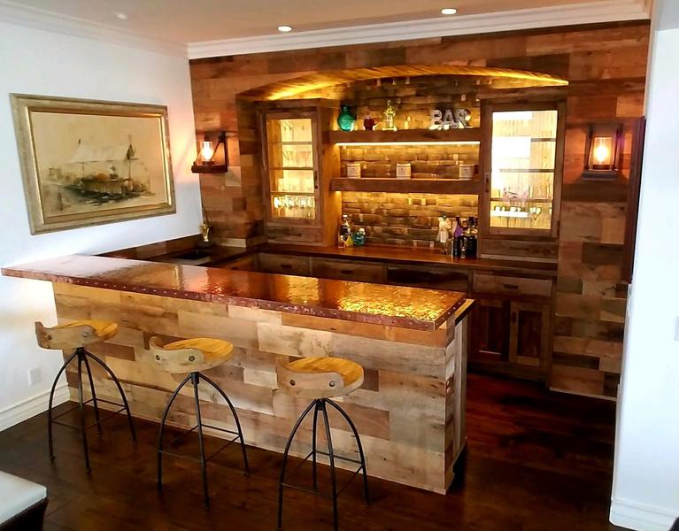 Rustic home bar with wood paneling, stone counter, and three stools in a warmly lit room