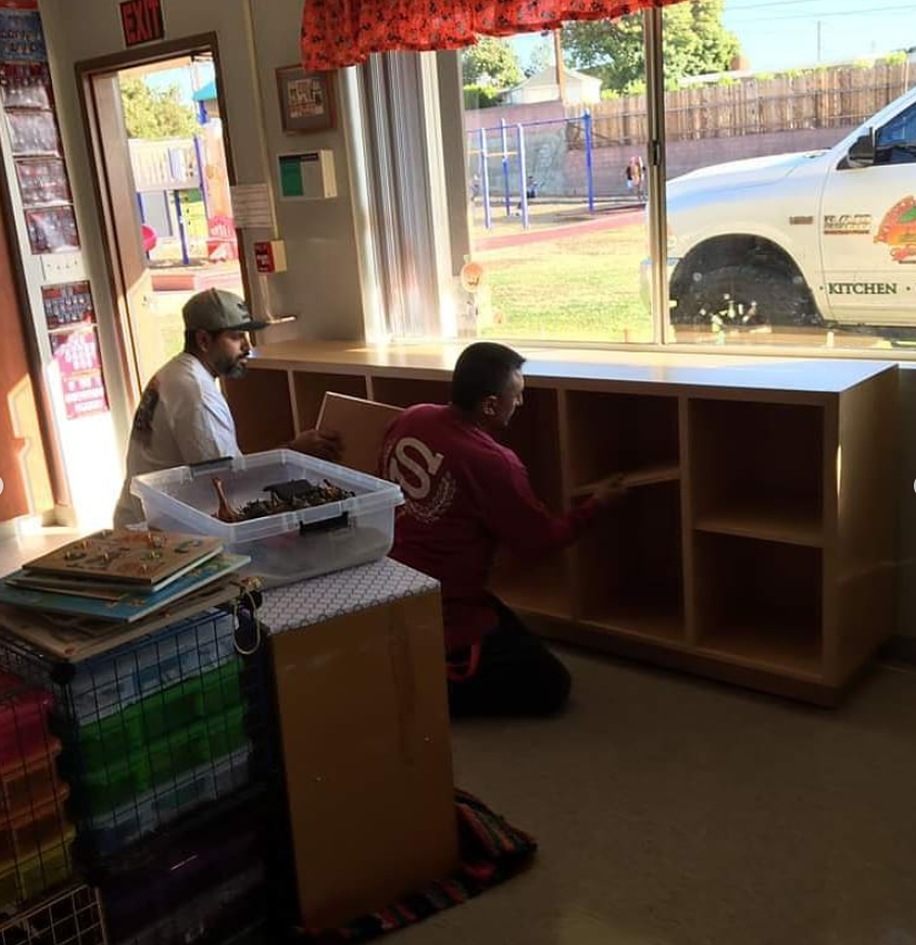Two people inside a shop, one kneeling by cubby shelves near a bright front window.