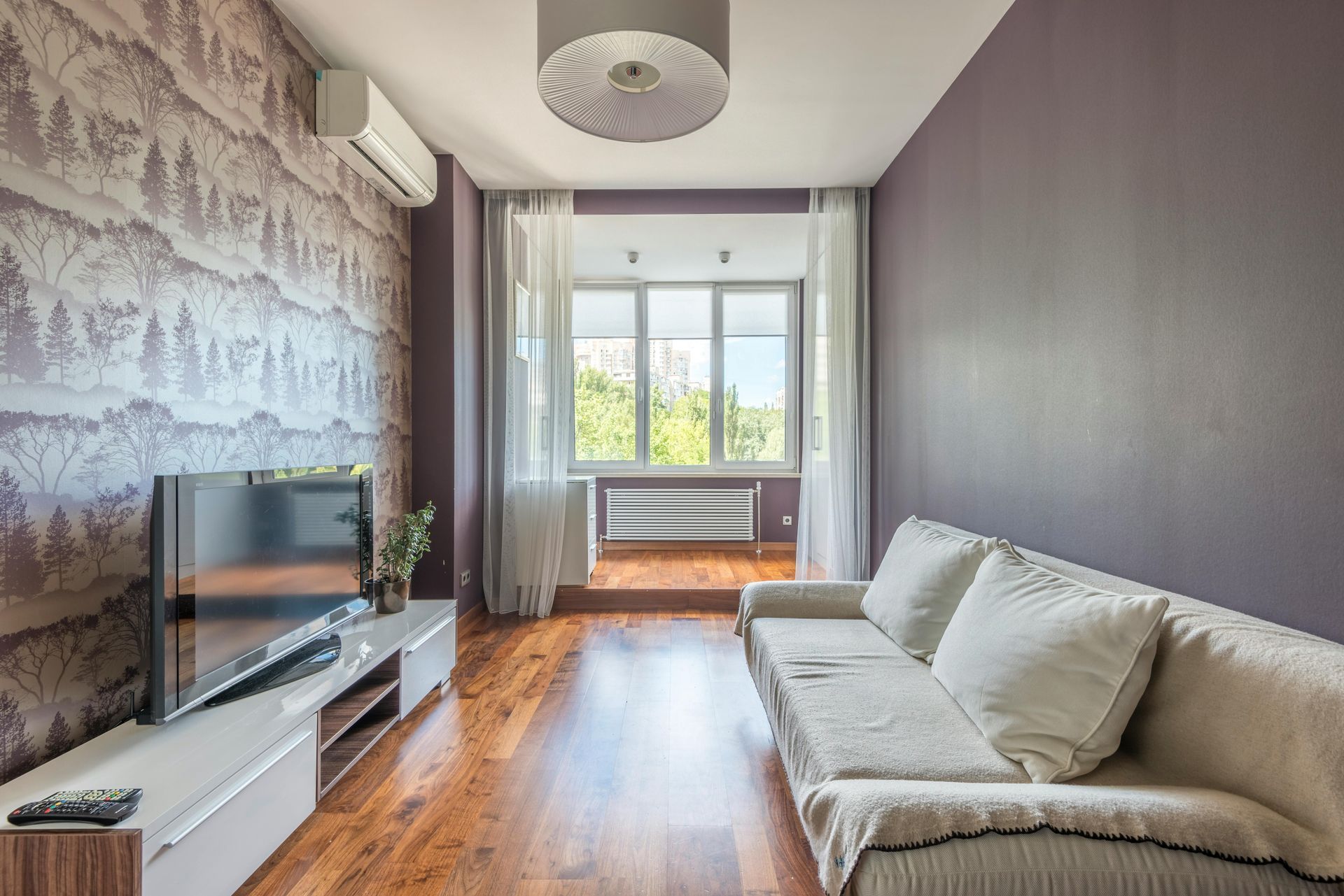 Living room with wood floors, couch, TV, patterned wallpaper, and window with natural light.