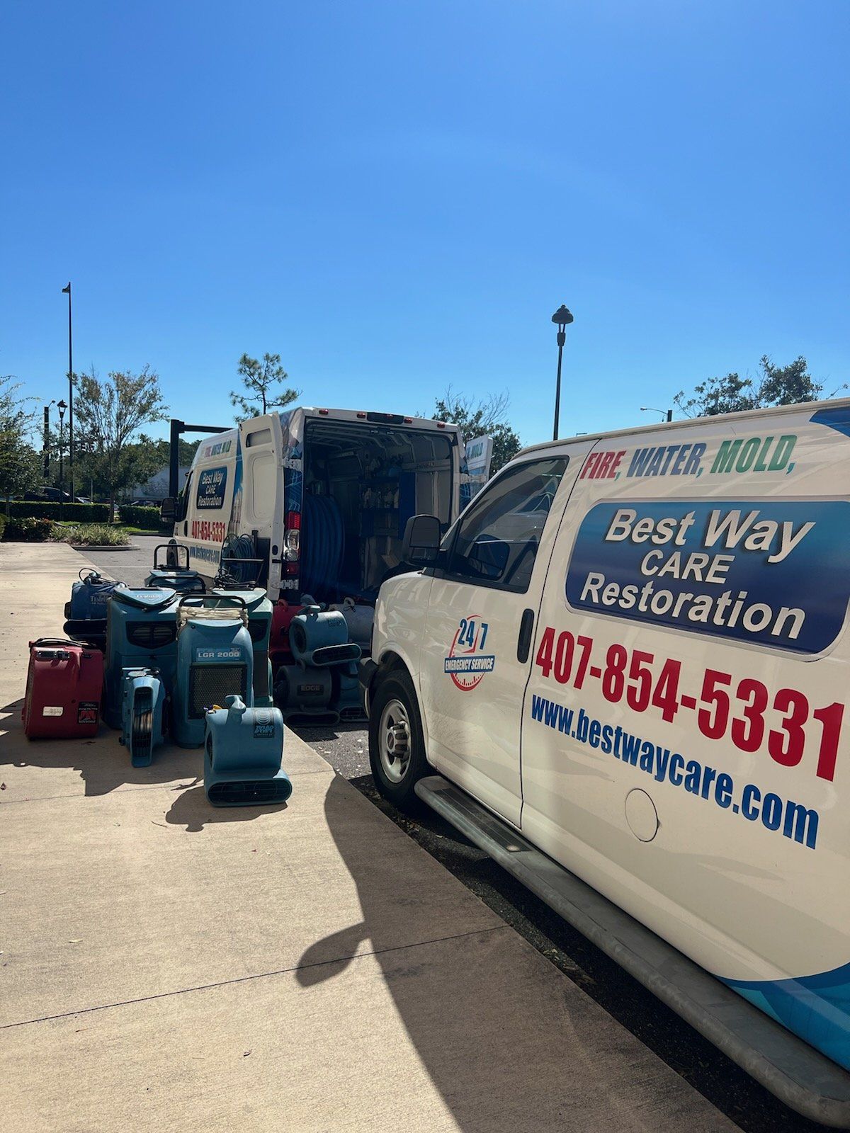Water damage restoration vans and equipment. White vans with logos, blue sky, concrete ground, and restoration tools.