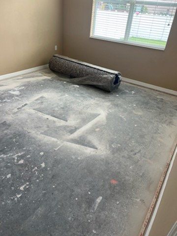 Bare concrete floor covered in dust, rolled-up rug, window with blinds.