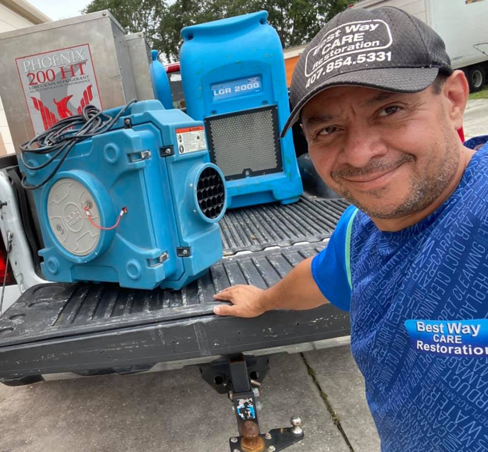 Man in truck bed with blue restoration equipment, smiling. Text on cap and shirt: 