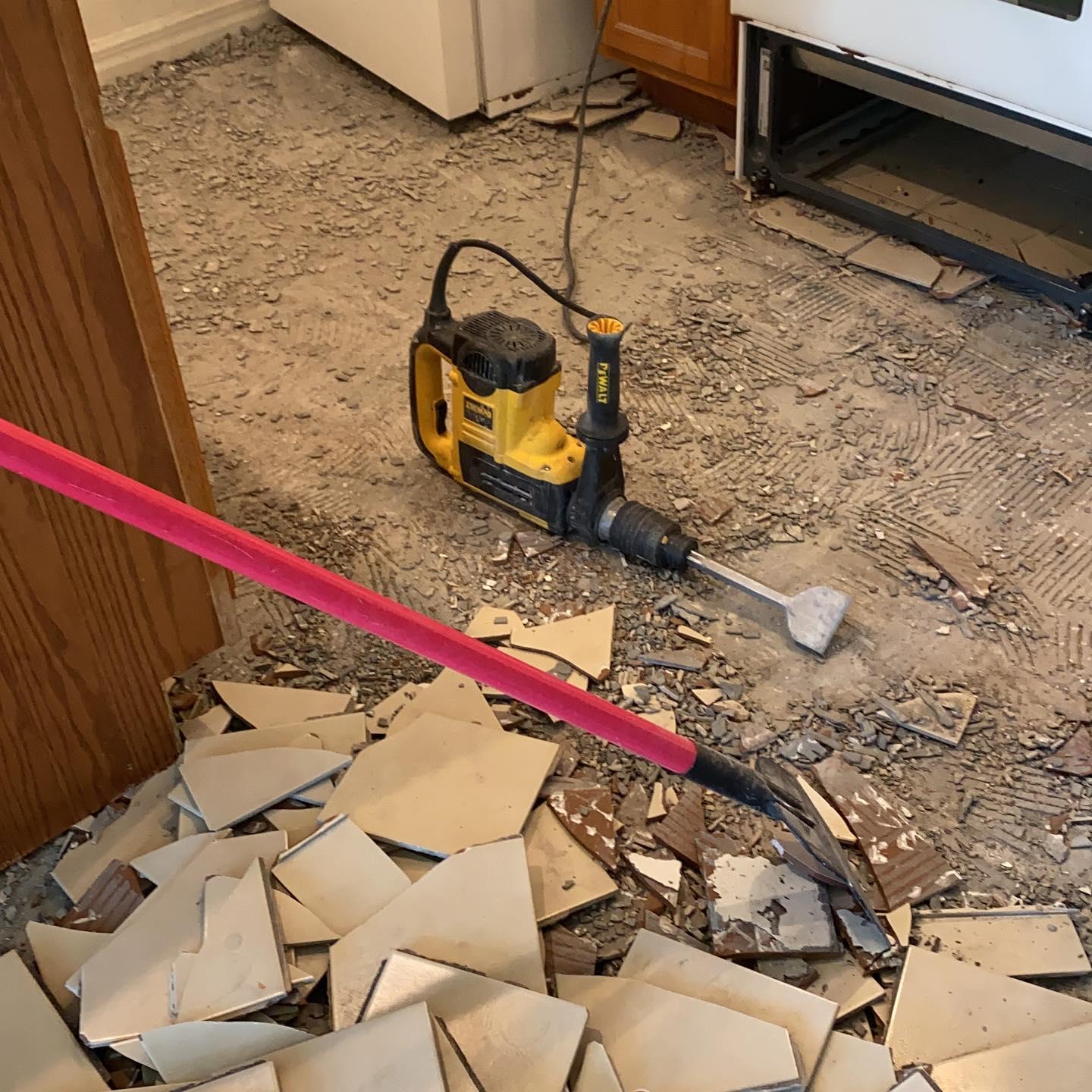 A kitchen floor being demolished with a jackhammer. Debris covers the floor; a pink handled shovel rests nearby.