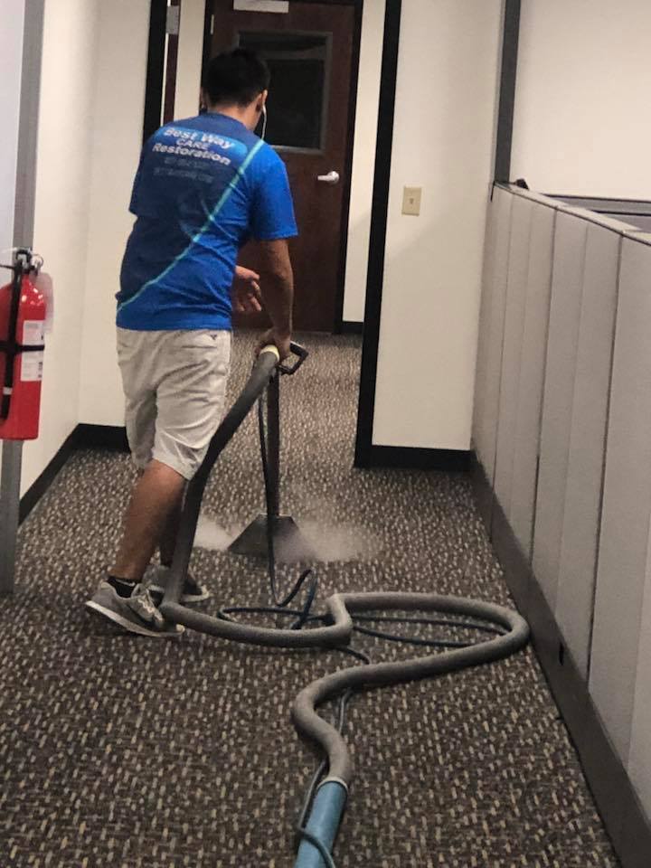 Person using a carpet cleaning machine in an office hallway; water spraying onto carpet.