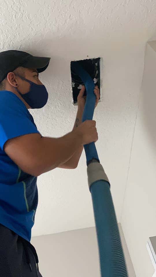 Person in blue shirt and mask vacuuming a ceiling vent opening with a blue hose.