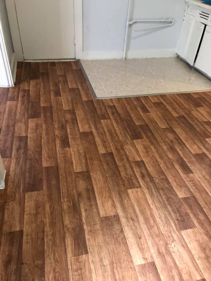 Brown wood-look flooring transitions to light beige flooring, near a white door, wall and kitchen appliance.