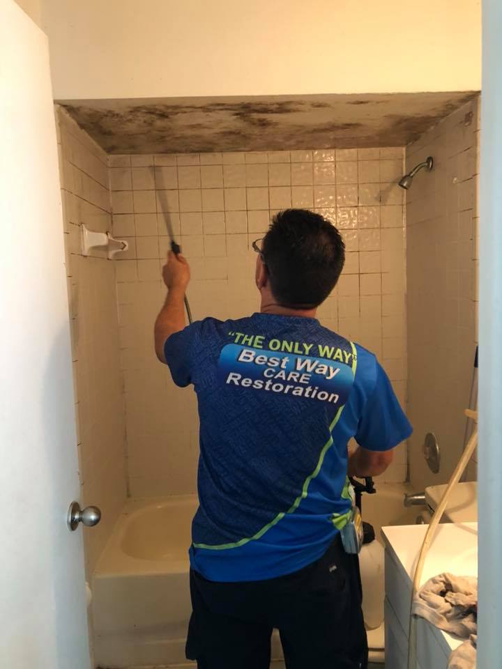 Person sprays a shower covered in mold, inside a bathroom, wearing a company shirt.