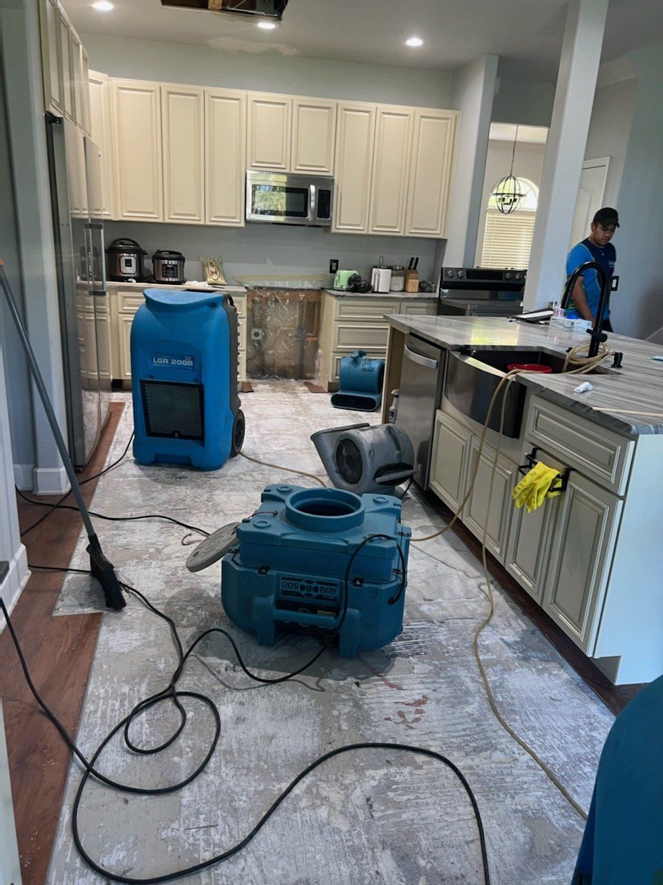 Kitchen with flood damage, drying equipment, and a person working. Cabinets, appliances, and floor are visible.