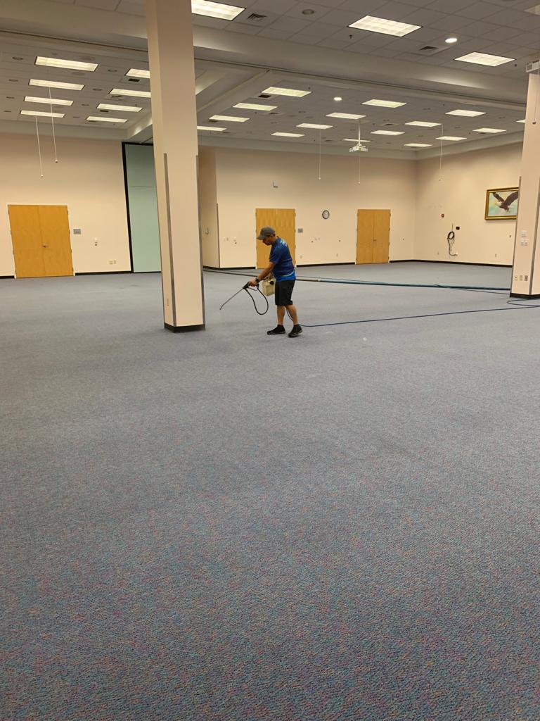 Man spraying cleaner on blue carpet in a large, empty room with white pillars and doors.