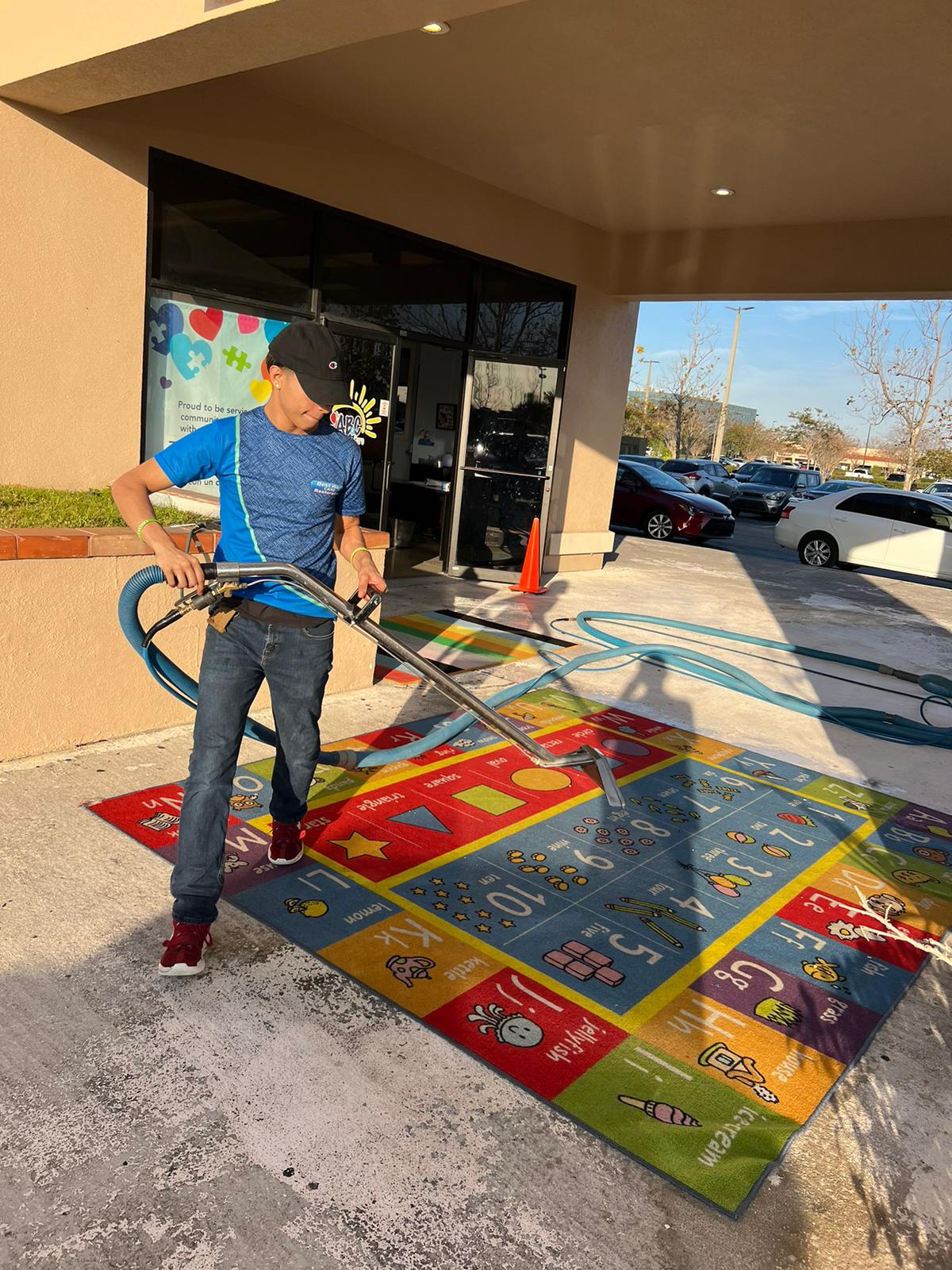 Man cleaning a colorful rug outside a building with a vacuum, sunlight, cars in the background.