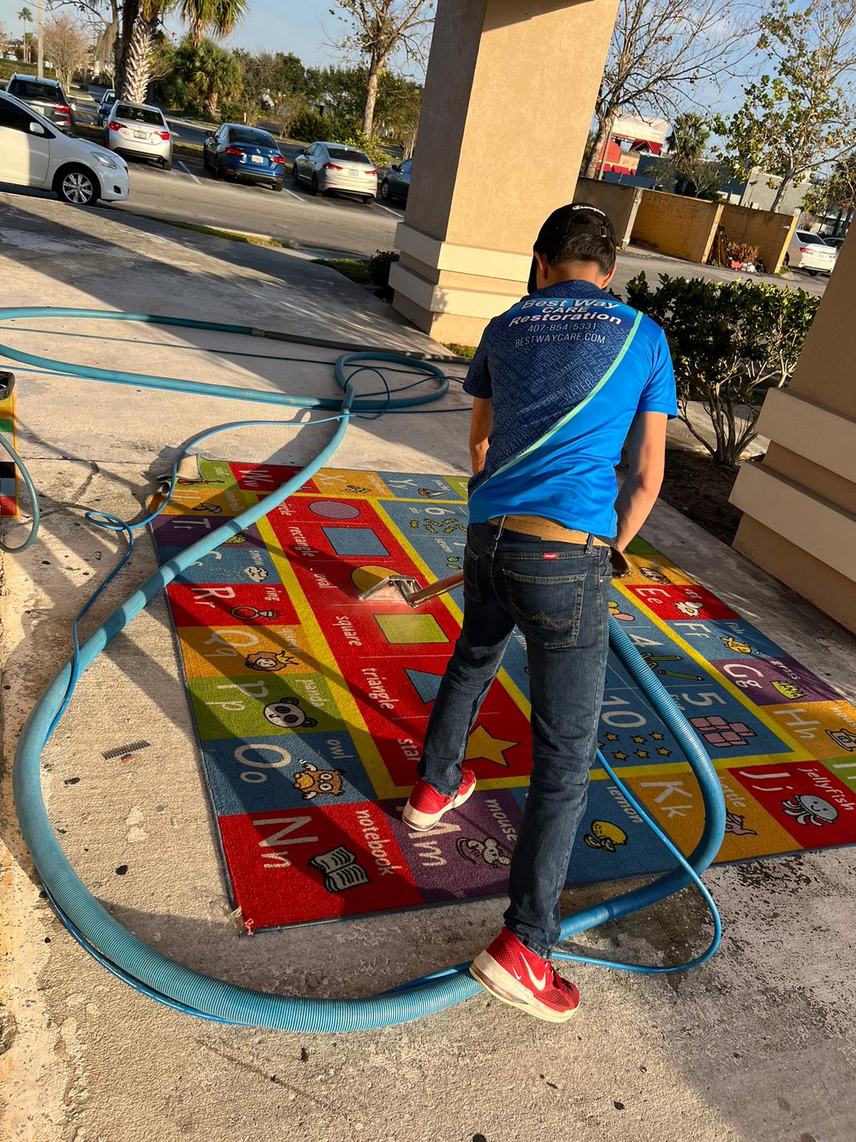 Man in blue shirt and jeans washes a colorful rug outdoors.