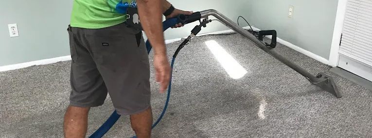 A technician in a green shirt uses a steam cleaning wand to clean gray carpet in a brightly lit, empty room.