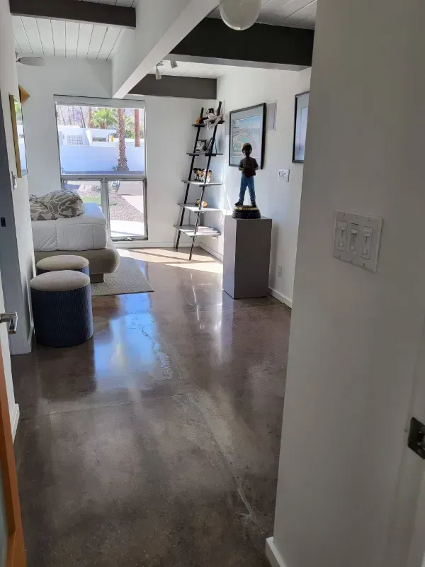 A sunlit room with polished concrete floors, a bed, two stools, a ladder shelf, and a statue on a pedestal.