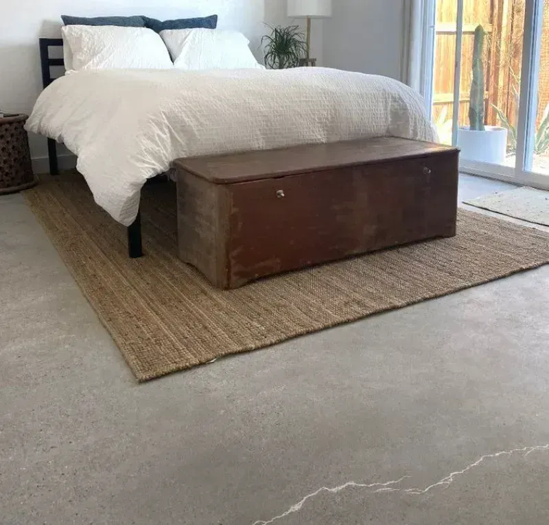 A bedroom with a white-covered bed and a dark wood storage trunk at its foot, set on a jute rug over a concrete floor.