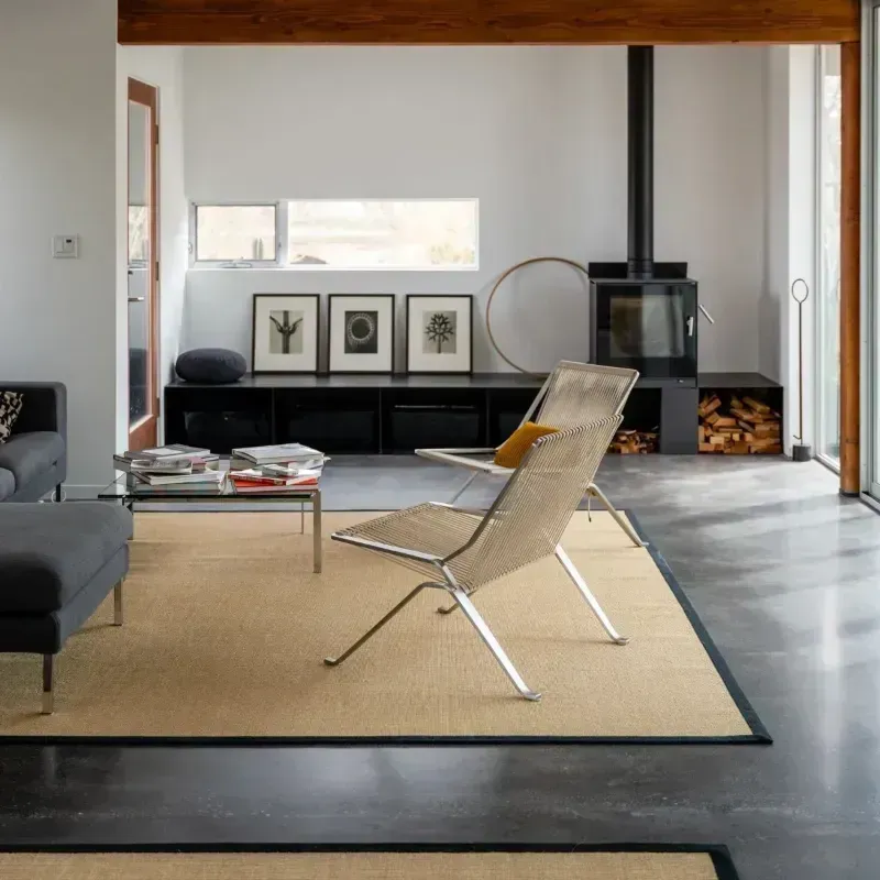 A minimalist living room with concrete floors, a tan area rug, two metal-framed chairs, and a fireplace with firewood.