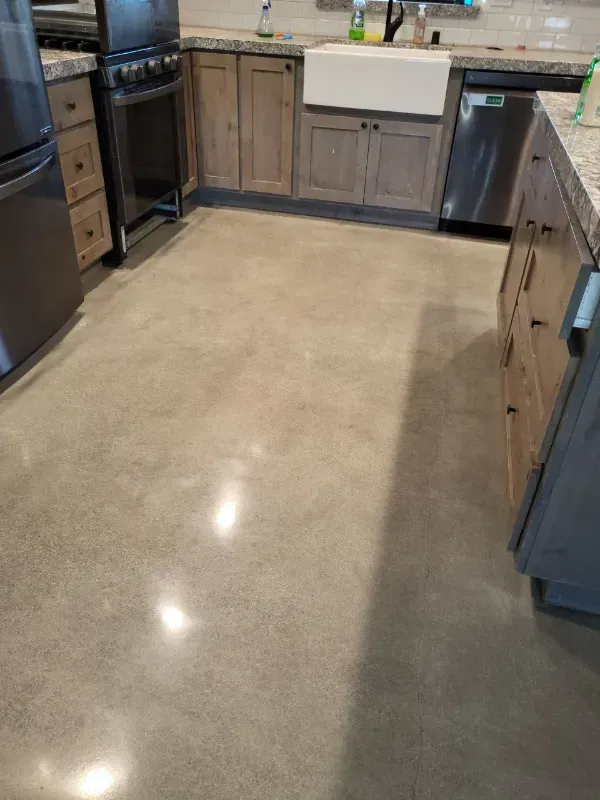 Polished concrete flooring in a kitchen with wooden cabinets, a white farmhouse sink, and stainless steel appliances.
