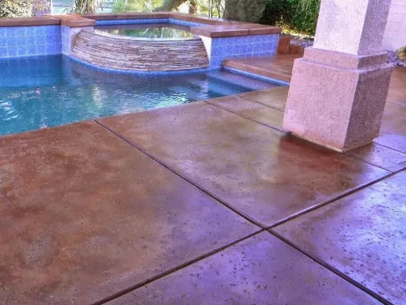 A stained, brown concrete pool deck adjacent to a swimming pool and a raised hot tub with blue tiled accents.