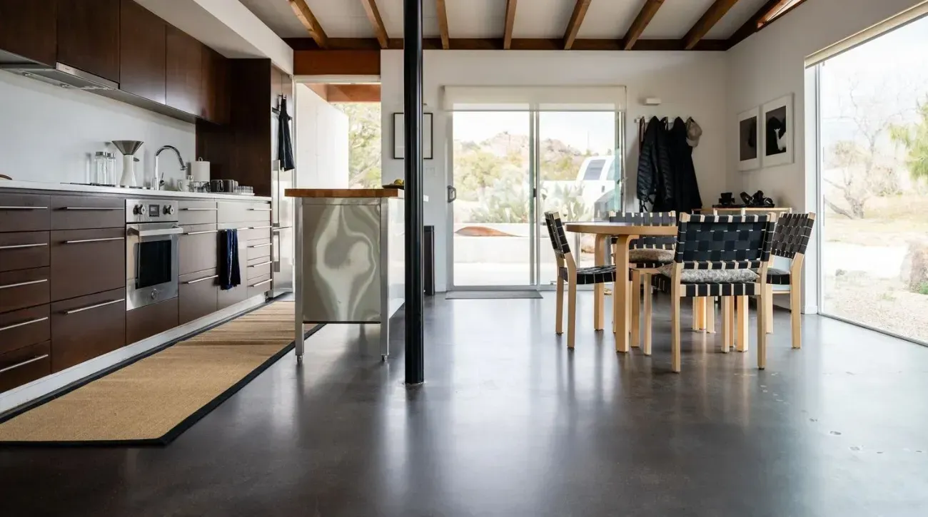 A modern kitchen and dining area with brown cabinets, a central island, and a dining set on a polished concrete floor.