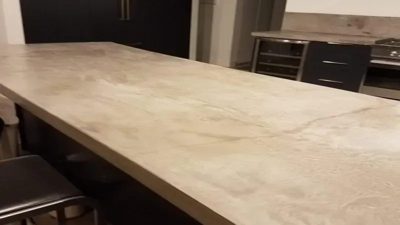 A long, light-colored, textured kitchen island countertop in a modern kitchen with black cabinets and bar stools.