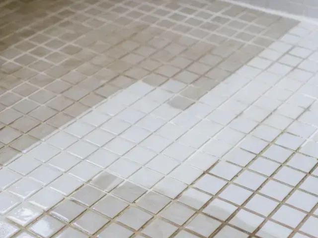 Close-up of a tiled shower floor. Light gray and white square tiles show cleaning progress.