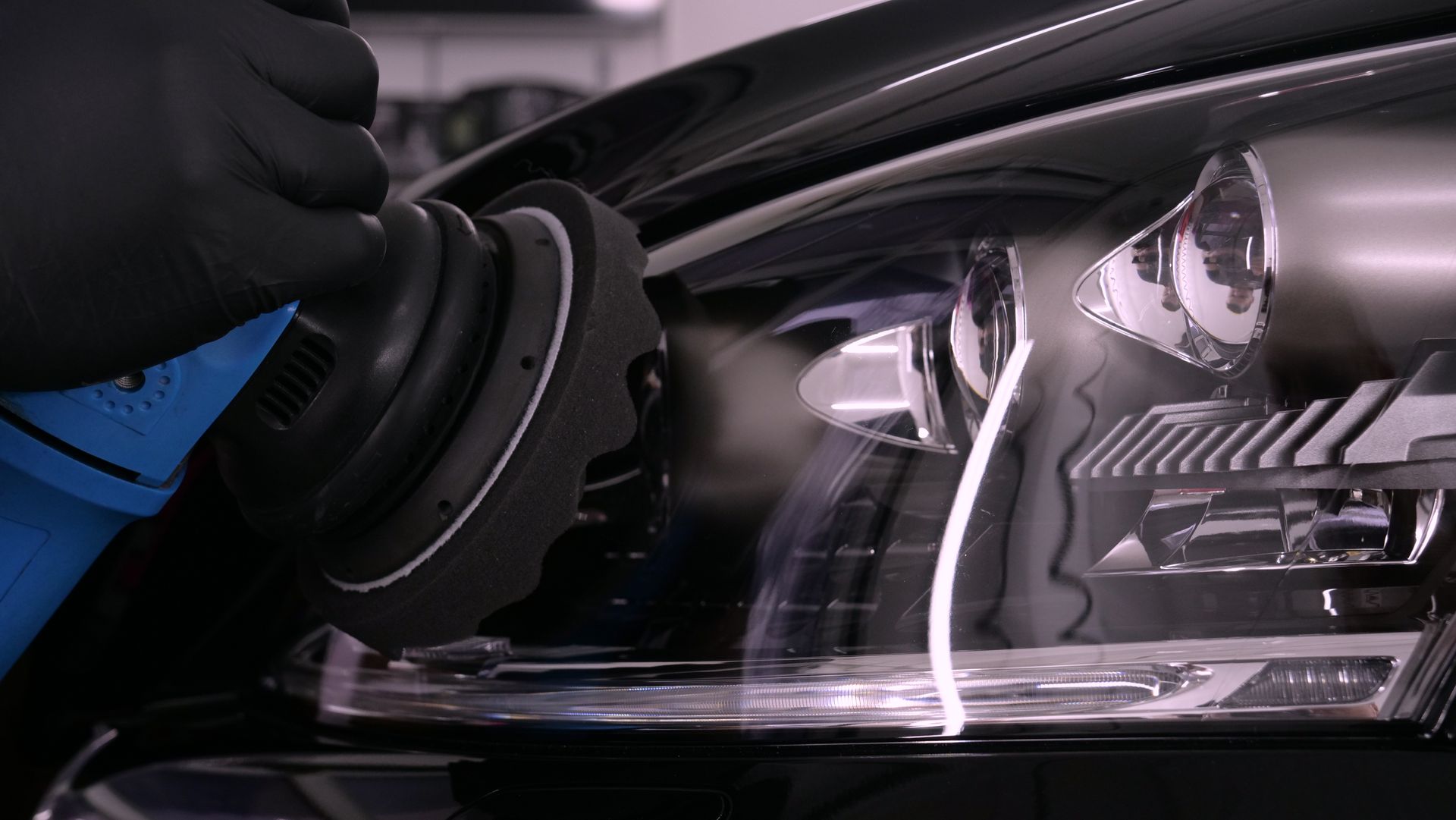 Black-gloved hand using a polishing tool on a car's headlight, detail shot, setting is inside.