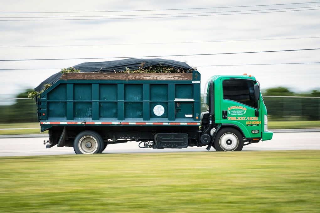 Green garbage truck driving on a road, carrying debris.