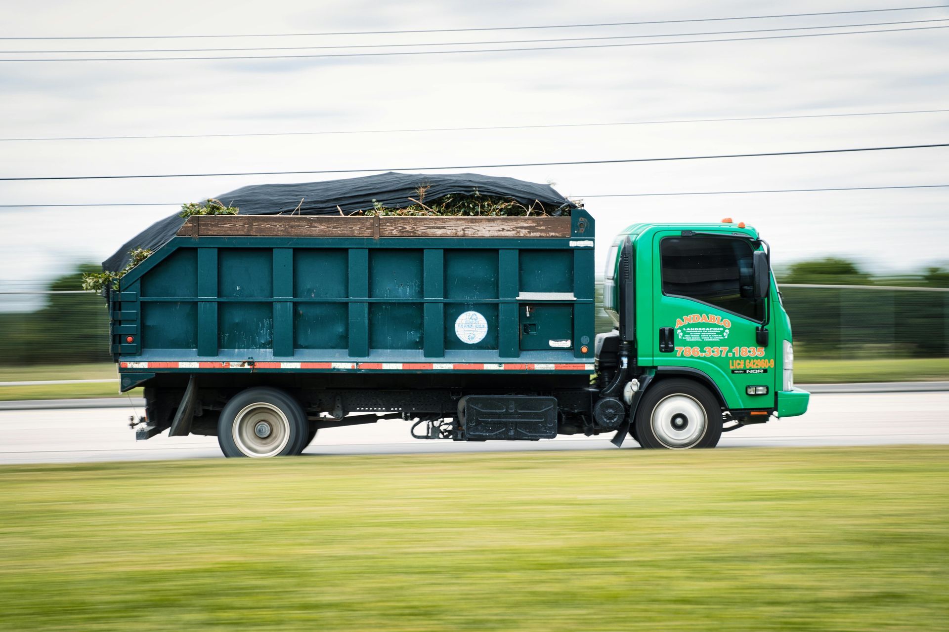 Green garbage truck driving on a road, carrying debris.