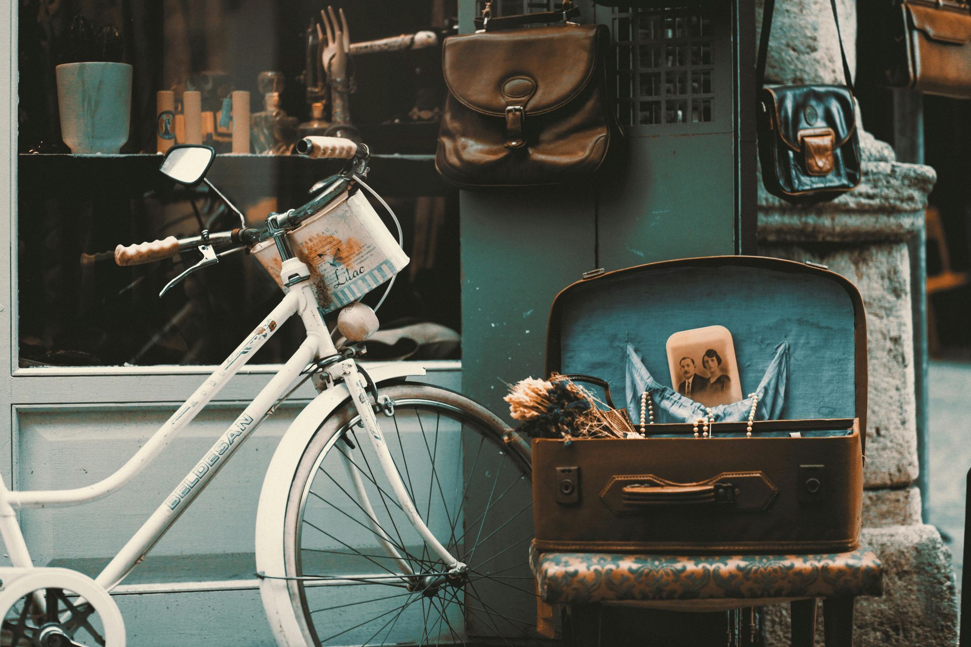 White bicycle next to a vintage suitcase, with a shop window display of bags and trinkets.