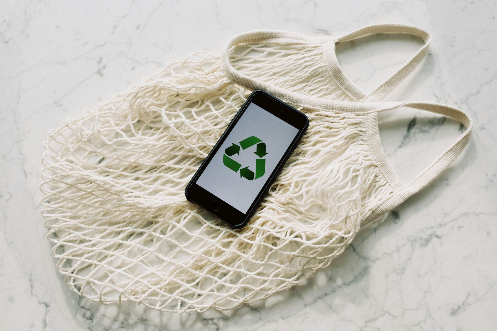 Smartphone with recycle symbol on screen rests inside a reusable mesh bag on a marble surface.