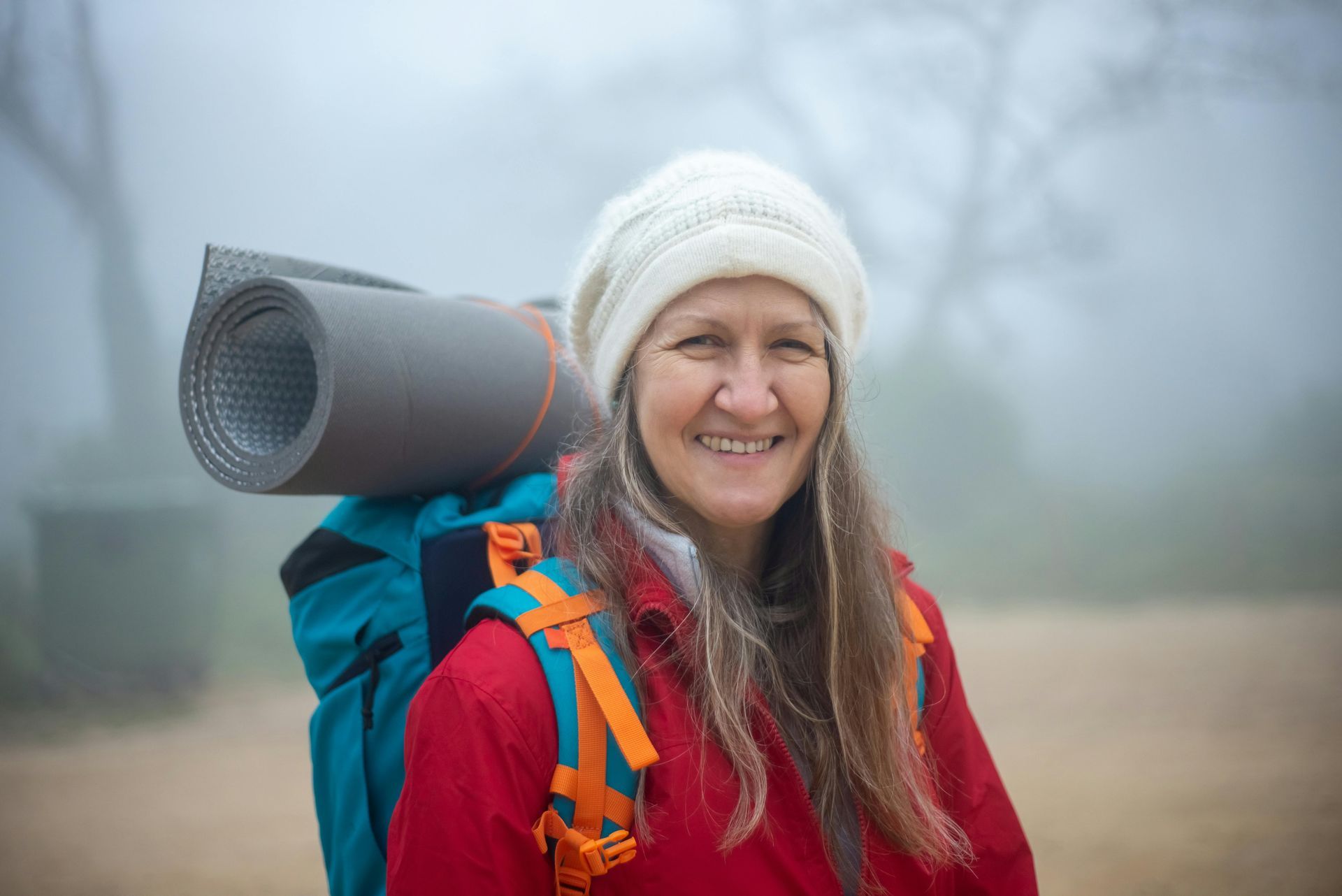 Woman with backpack, smiling, in a foggy outdoor setting, wearing a red jacket and white hat.