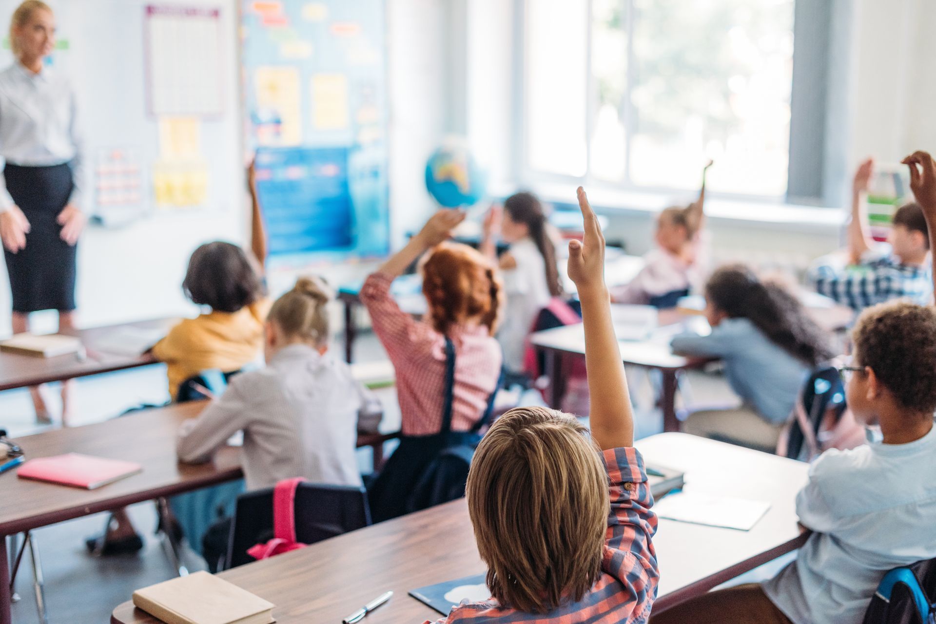 Students in a classroom raising hands while the teacher stands at the front near a whiteboard Students in a classroom raising hands while the teacher stands at the front near a whiteboard