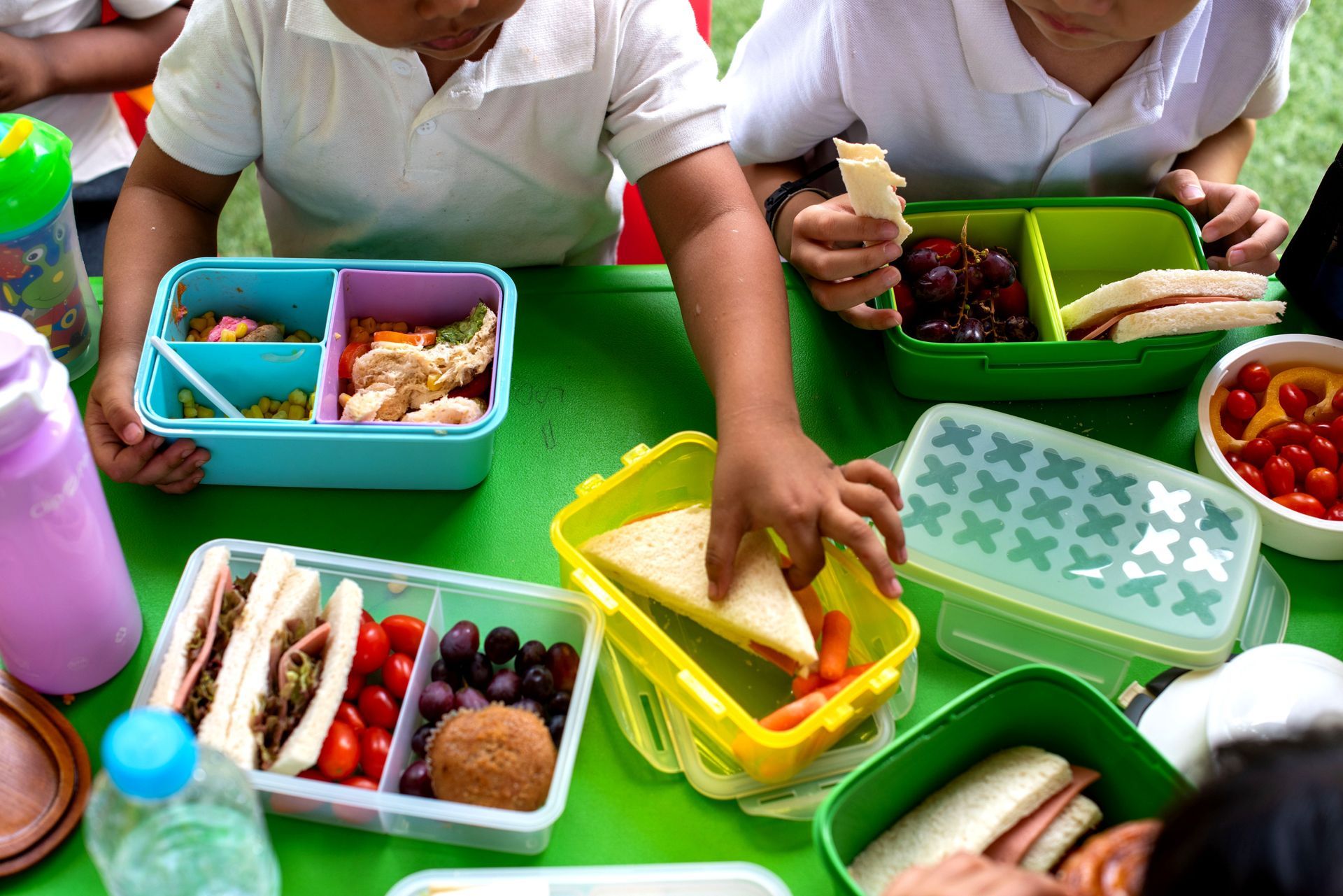 Children reaching for assorted food containers on a green table outdoors Children reaching for assorted food containers on a green table outdoors