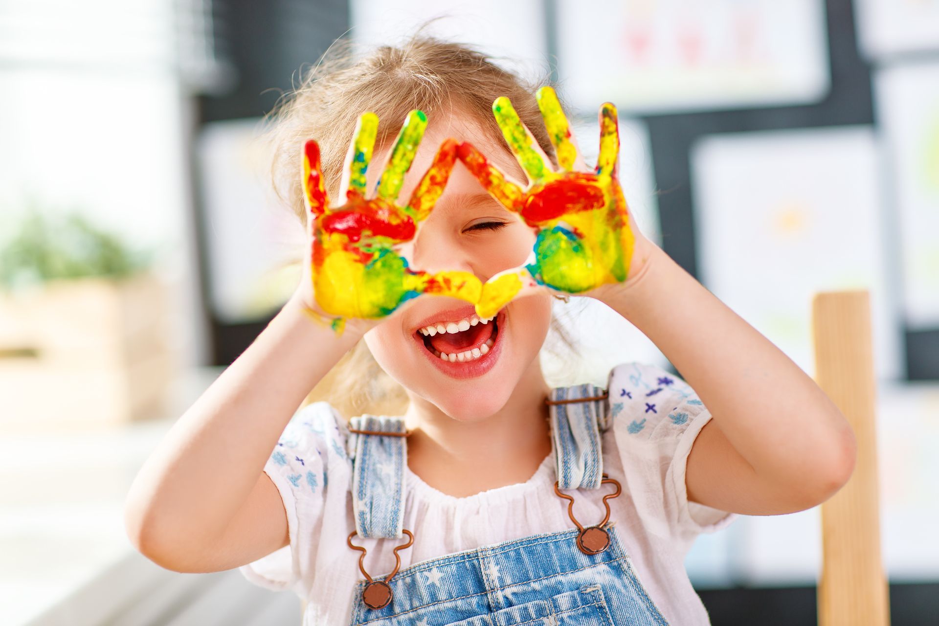 Child smiling with rainbow-painted hands held over eyes, in a bright indoor setting Child smiling with rainbow-painted hands held over eyes, in a bright indoor setting