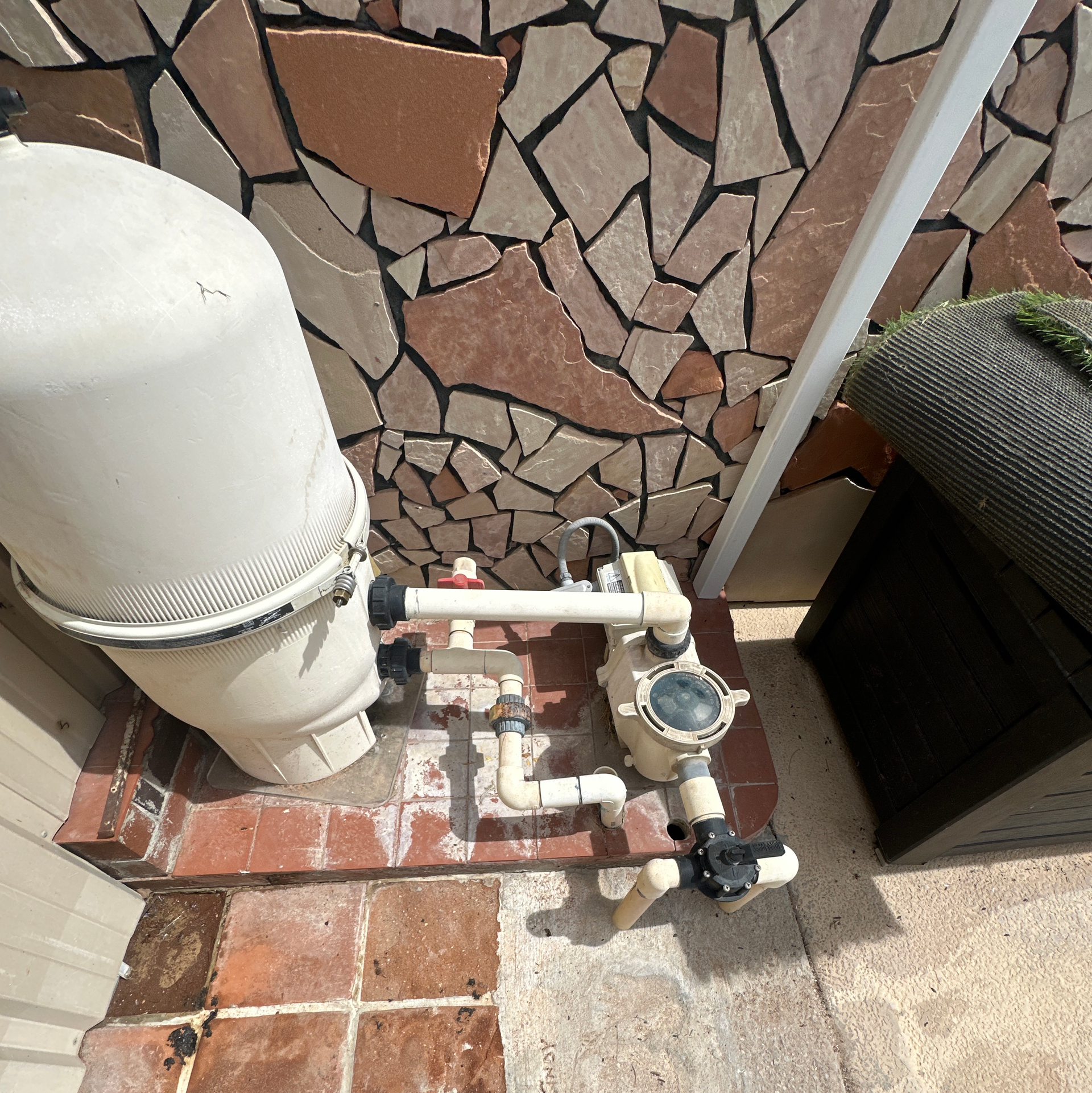 Pool plumbing and filter equipment beside a stone wall and patio tiles.