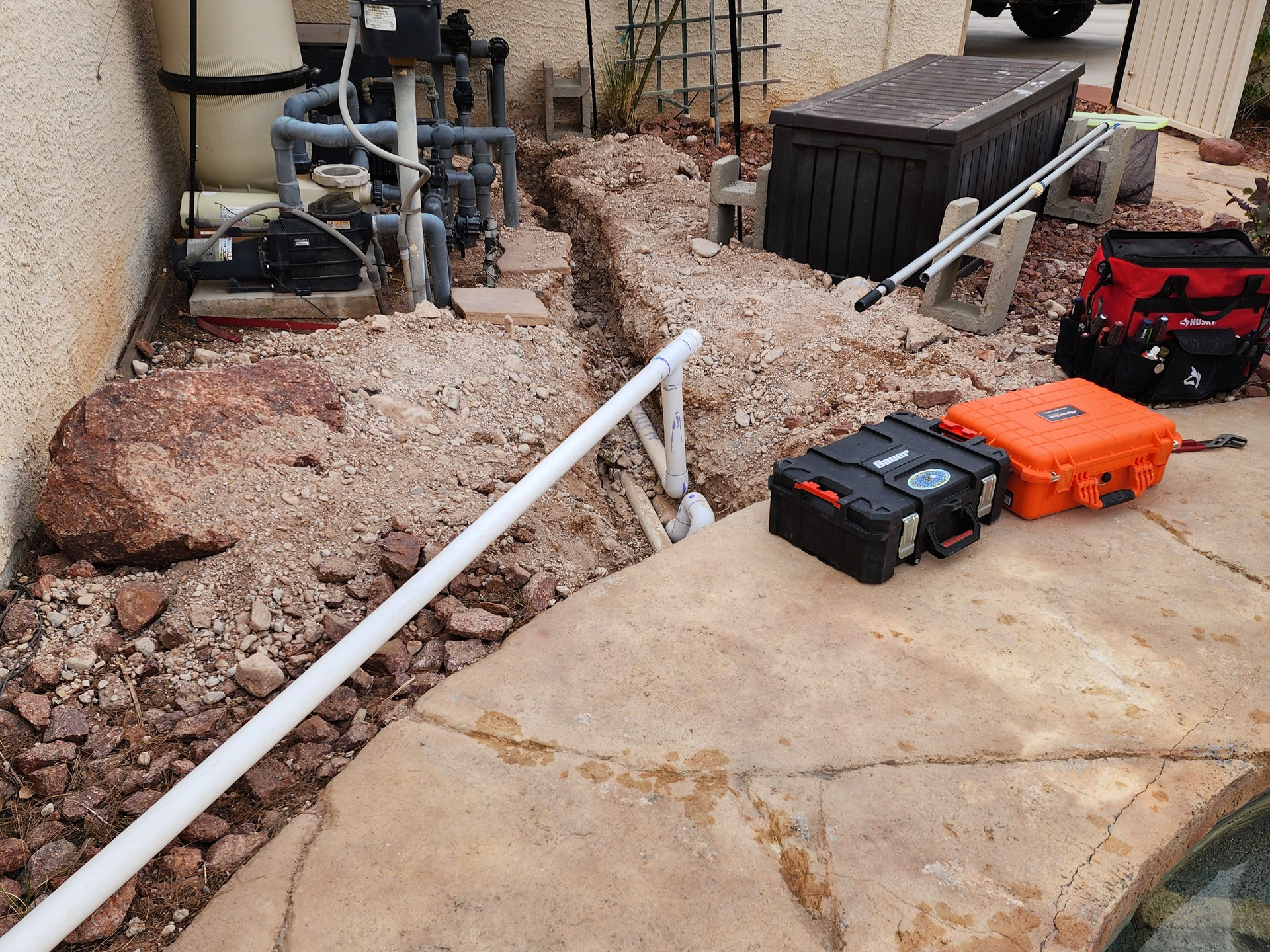 Backyard with pool equipment, gravel trench, white PVC pipe, and two toolboxes on a concrete patio