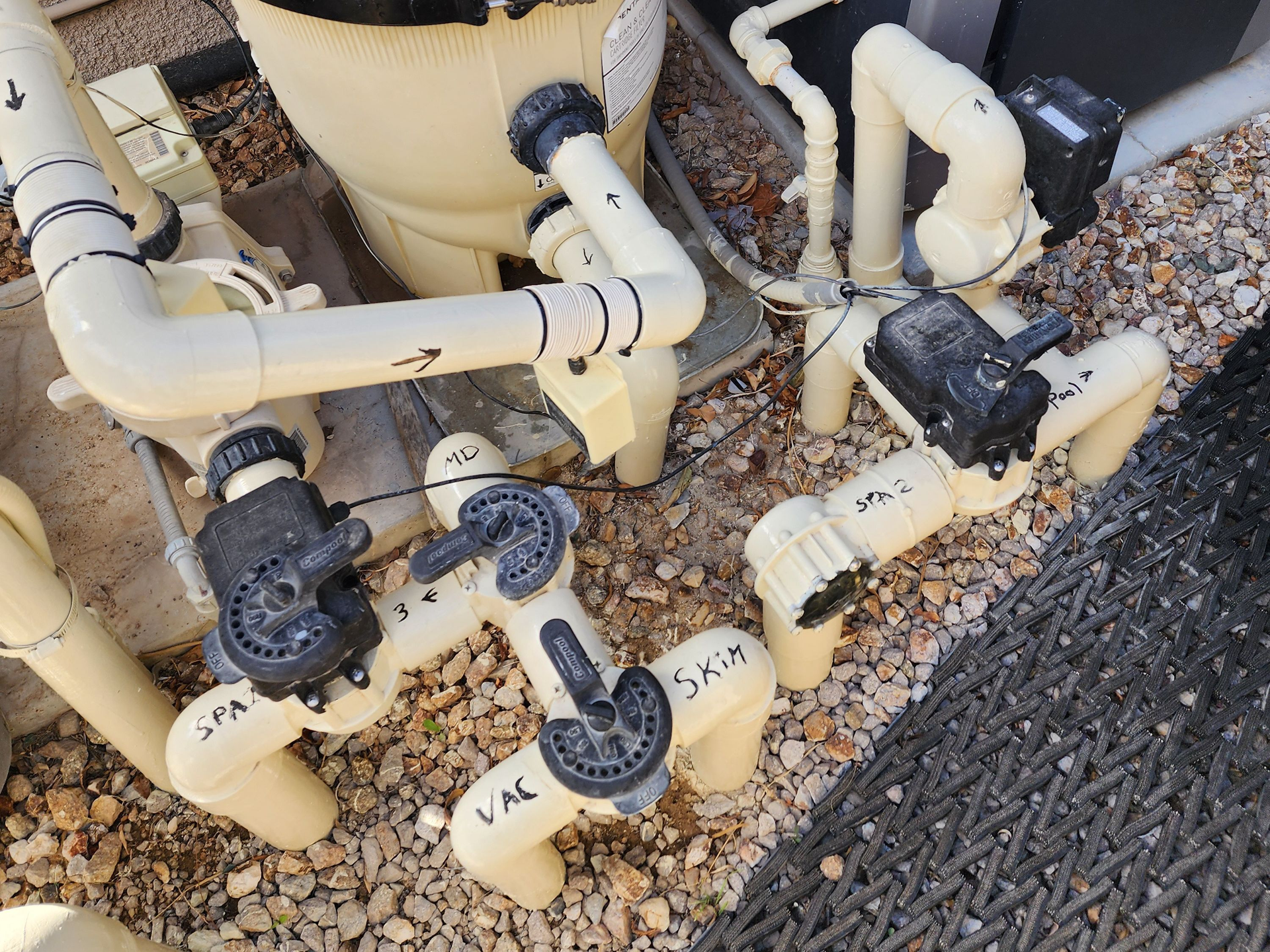 Beige pool plumbing pipes with black valves and fittings on a gravel bed.