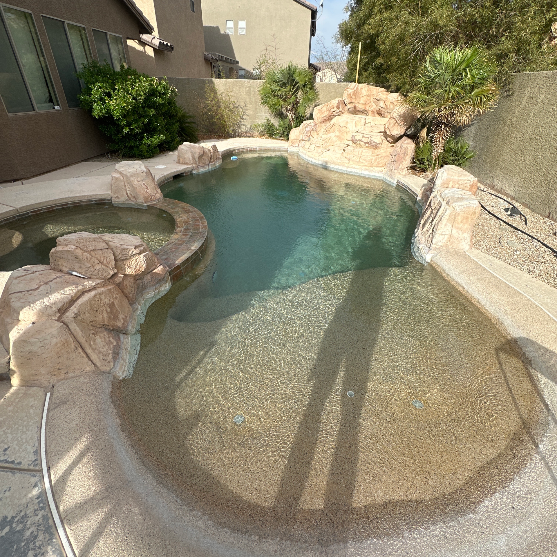 Backyard pool with rock-edge spa and clear water, surrounded by stucco walls and desert landscaping.