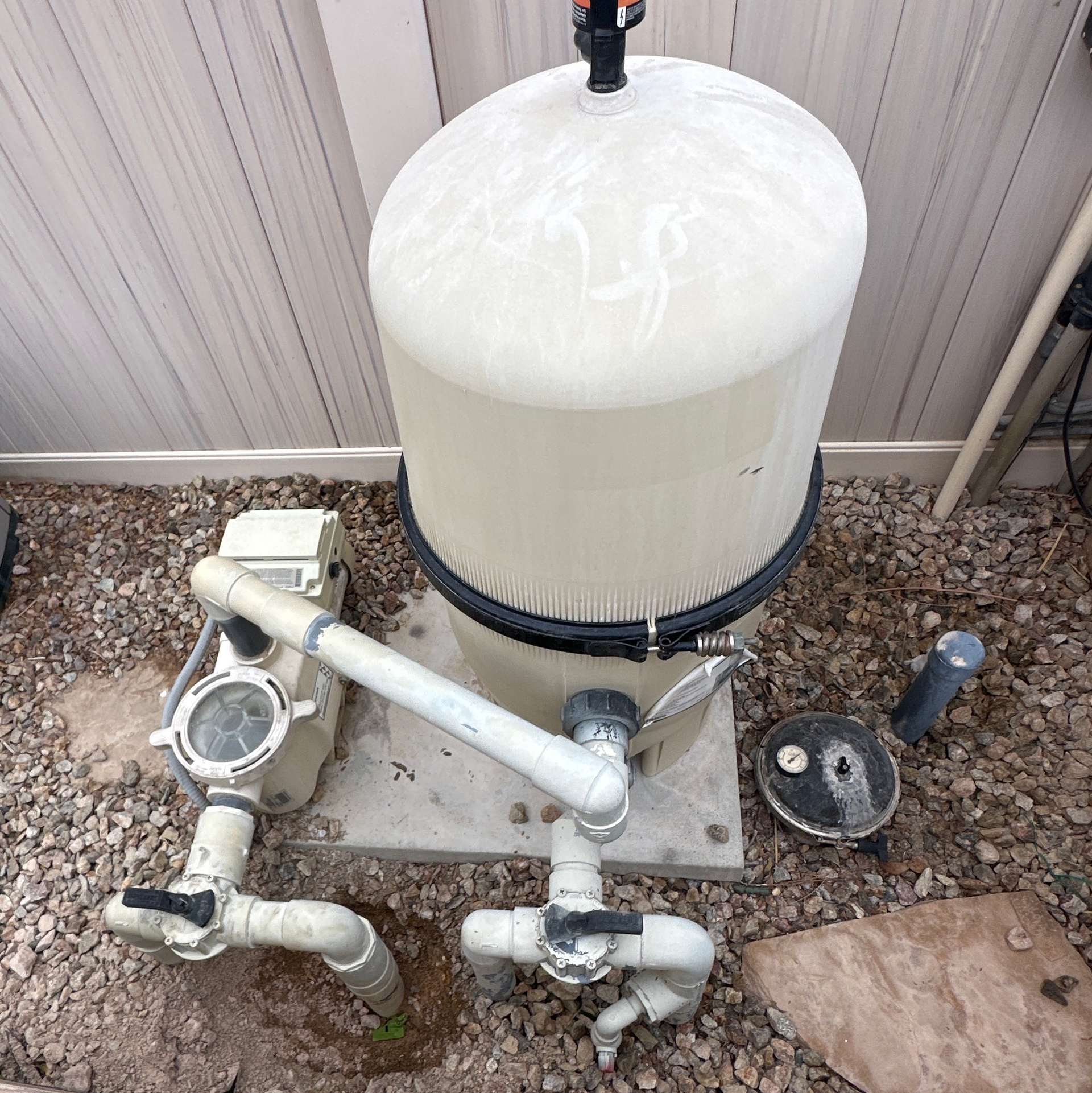 White cylindrical tank with pipes and valves on a gravel pad beside a building