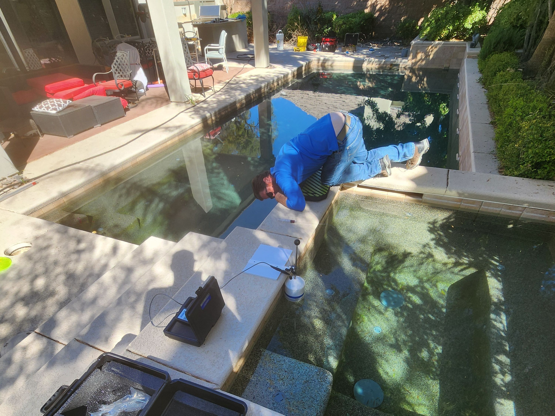 Person in a blue jacket crouches on a stone ledge beside a backyard pool or pond, reaching into the water.
