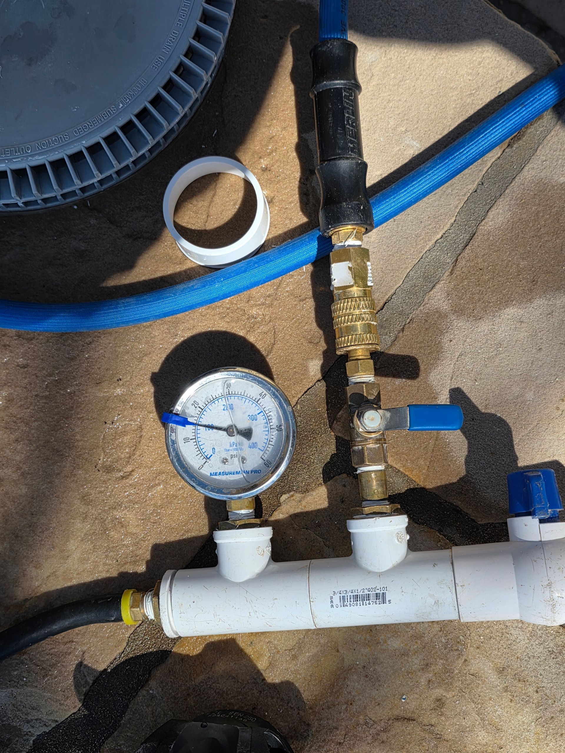Pressure gauge and brass valves on white PVC pipe with blue hoses and a wheel in the background.