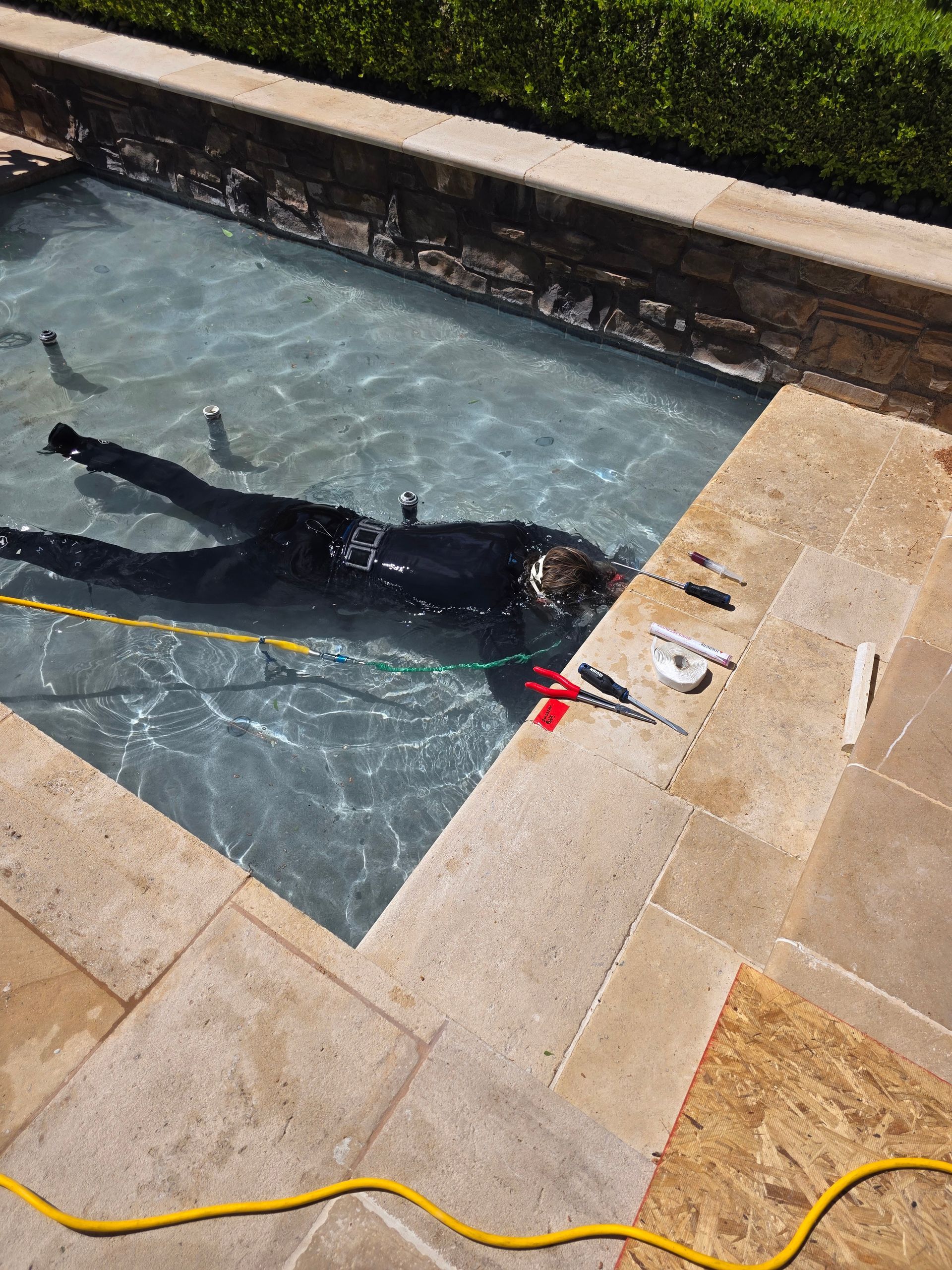 Black dog floating in a backyard pool beside tan stone decking and a yellow hose