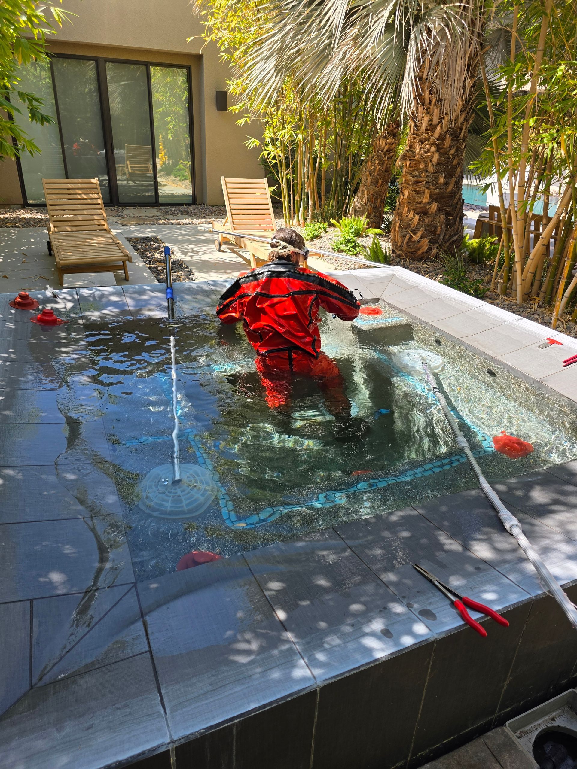 Person kneeling by a backyard pool, cleaning the water with a hose and pool vacuum