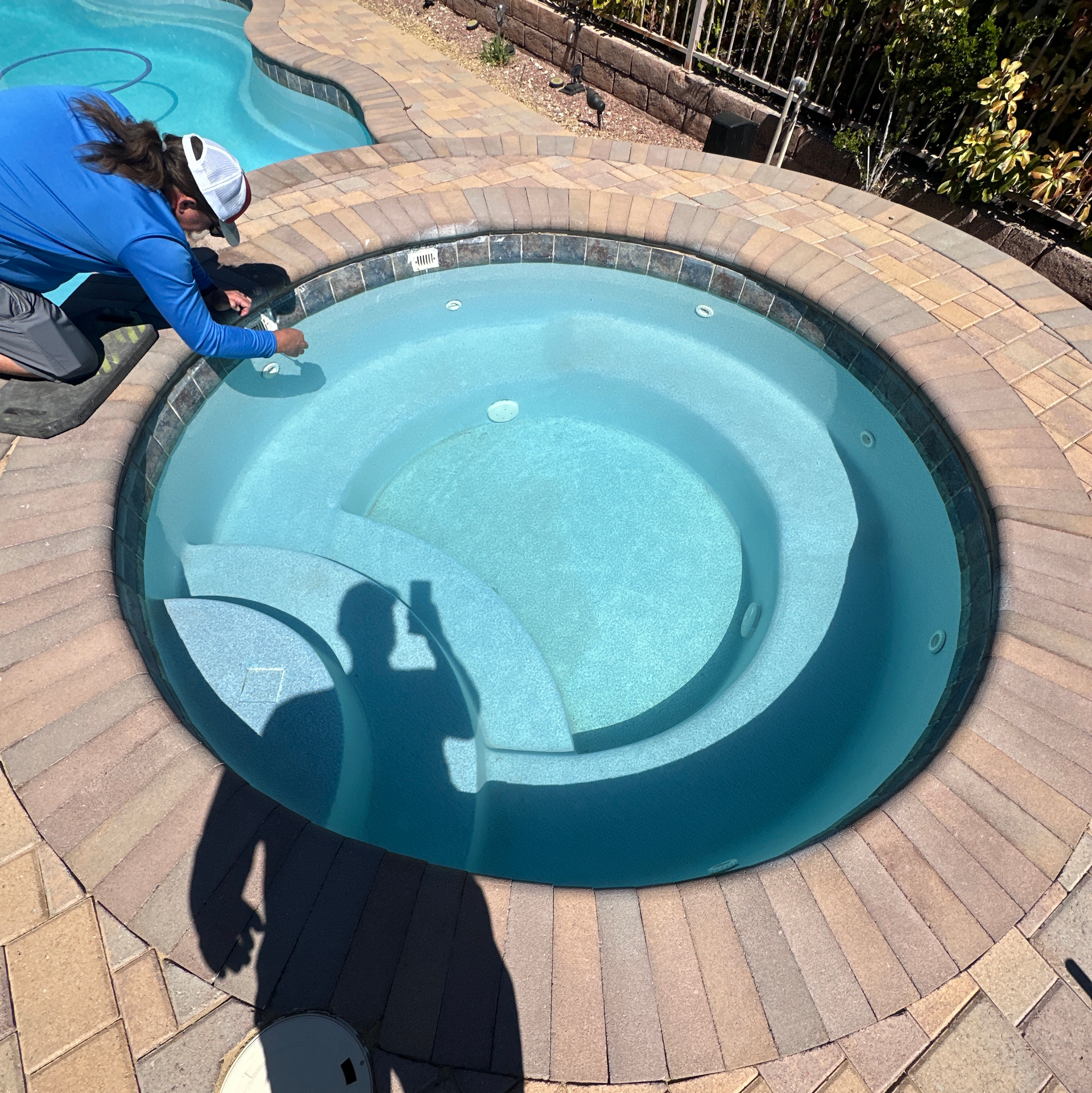Person kneeling beside a small backyard pool, cleaning the water with a tool in bright sunlight